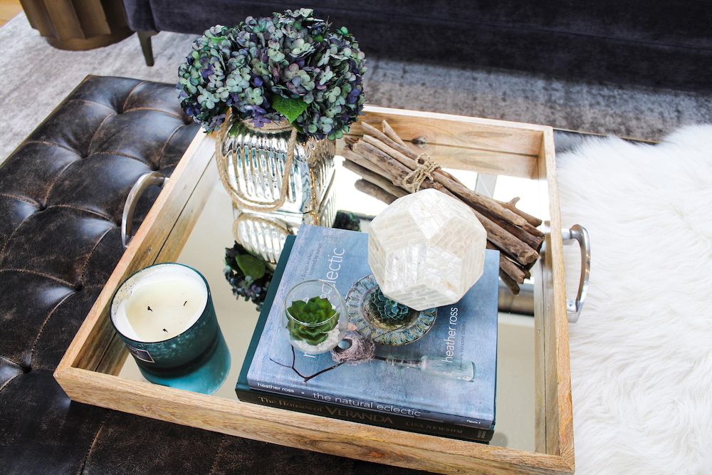 A styled wooden tray with metal handles sits atop a leather tufted ottoman.